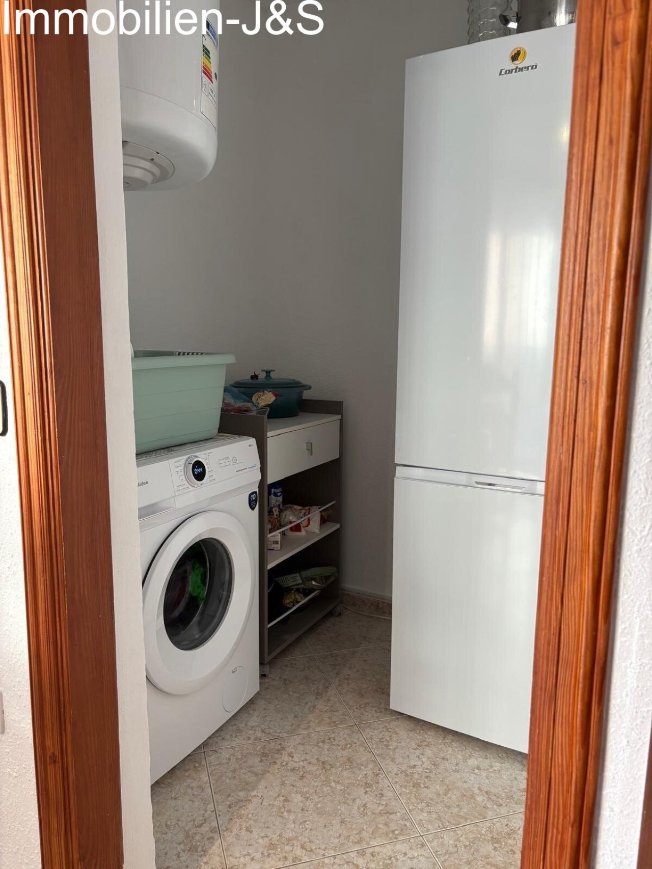 Praktischer Hauswirtschaftsraum direkt neben der Küche, ausgestattet mit Waschmaschine, Warmwasserboiler und zusätzlichem Platz für Kühlgeräte oder Vorräte.