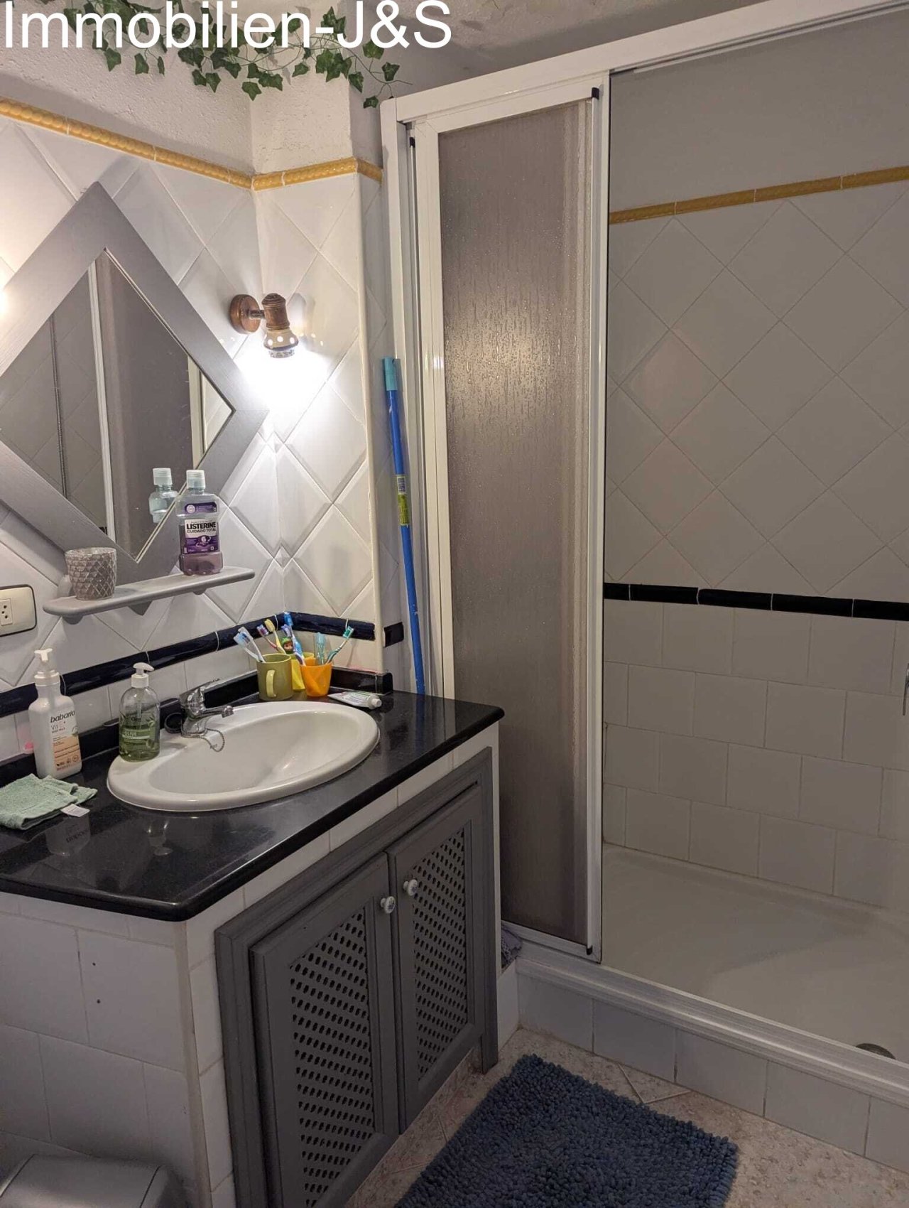 Das Badezimmer überzeugt durch ein zeitloses Design mit hellen Wandfliesen und dunklen Akzenten. Es ist funktional ausgestattet mit einer Duschtasse, einem Waschtisch mit praktischem Unterschrank sowie einem eleganten, rautenförmigen Spiegel.