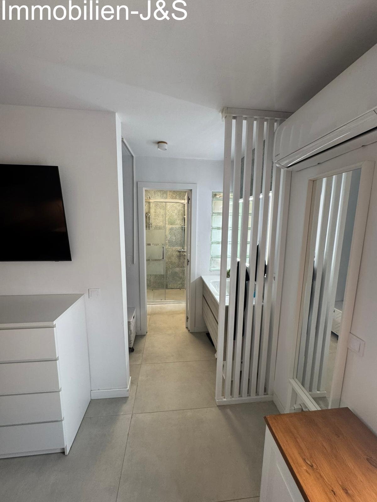 Direkter Zugang vom Schlafzimmer in das moderne En-Suite-Badezimmer. Ein stilvoller Raumtrenner bewahrt die Offenheit des Raumes, während das Bad mit einer großzügigen, ebenerdigen Dusche und hochwertiger Ausstattung überzeugt.