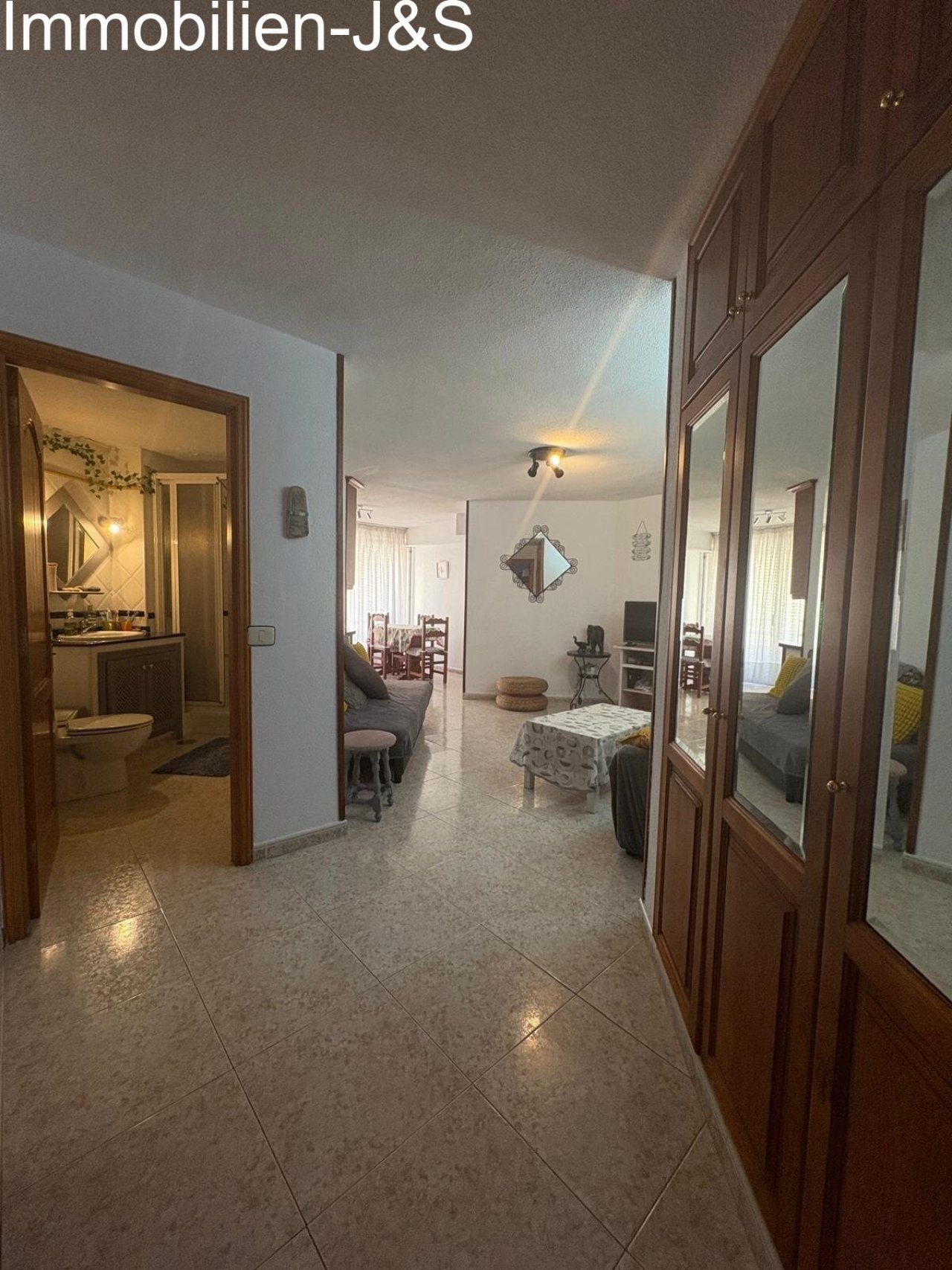 View from the entrance area into the brightly tiled hallway, past a spacious built-in wardrobe with mirrored doors, directly into the cozy living area.