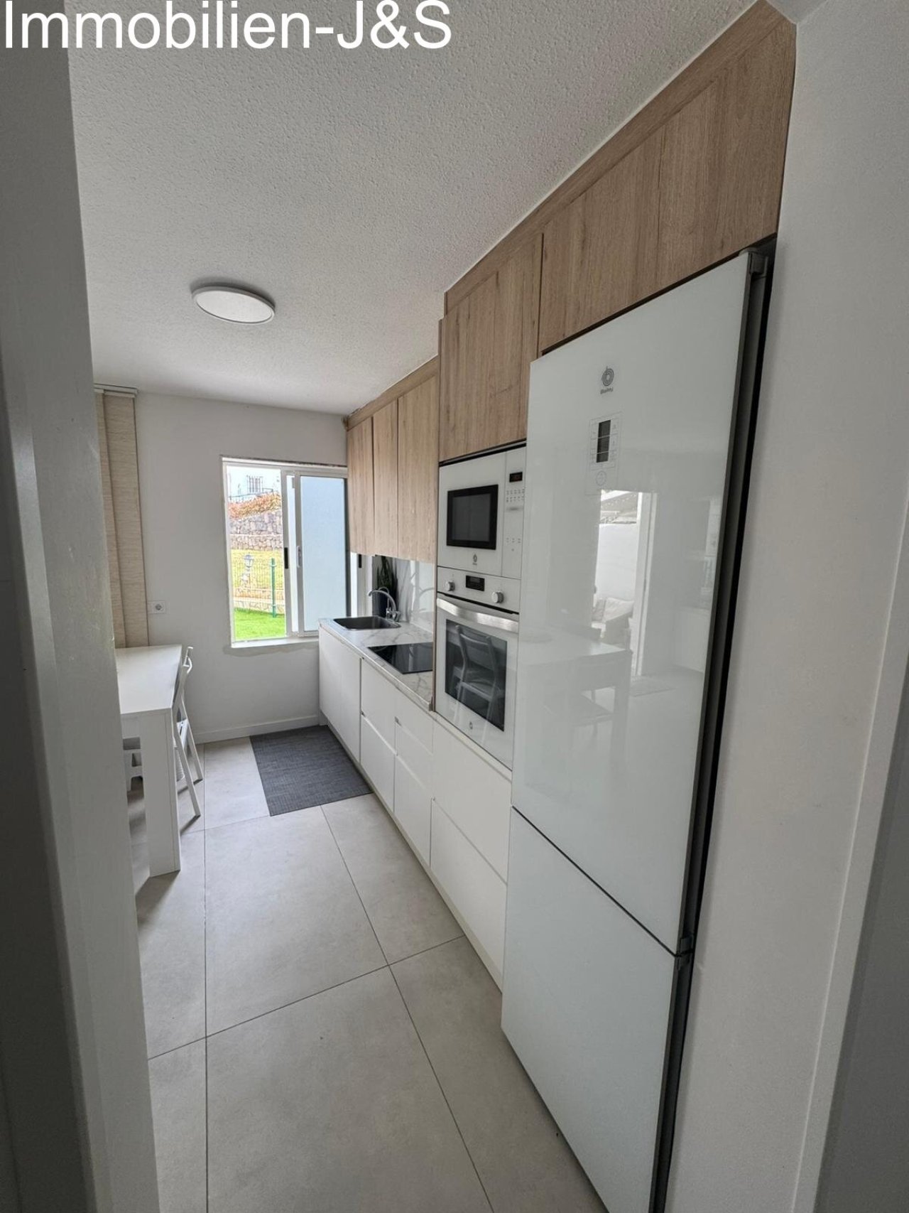 High-quality, handleless kitchen units in white with upper cabinets in a warm wood finish. Fully equipped with state-of-the-art appliances, it offers ample storage and a pleasant view through the adjacent window.