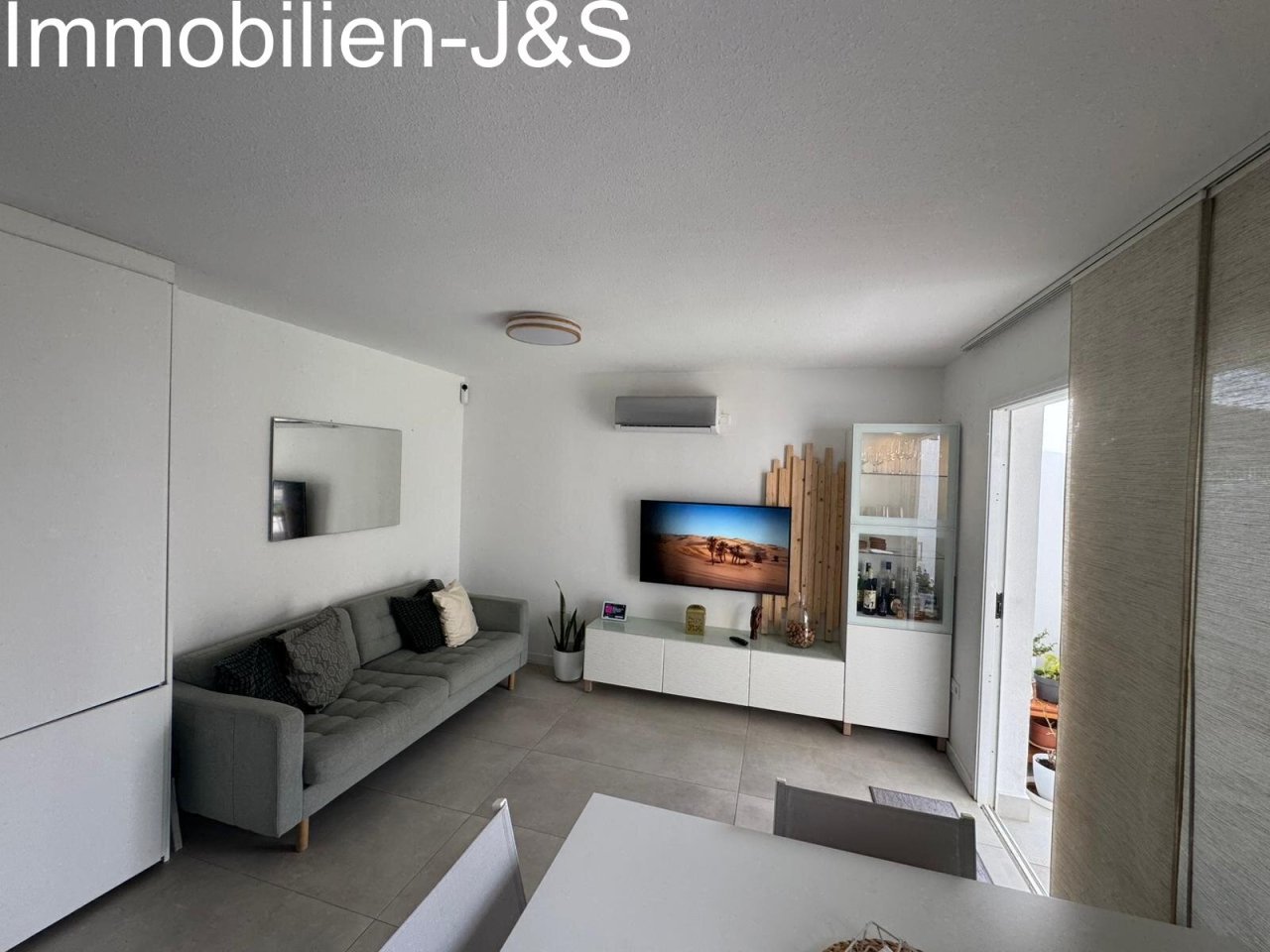 A bright, minimalist living area with clean lines. Featuring air conditioning, a comfortable sofa, and a stylish TV unit, this space offers contemporary comfort and direct outdoor access.